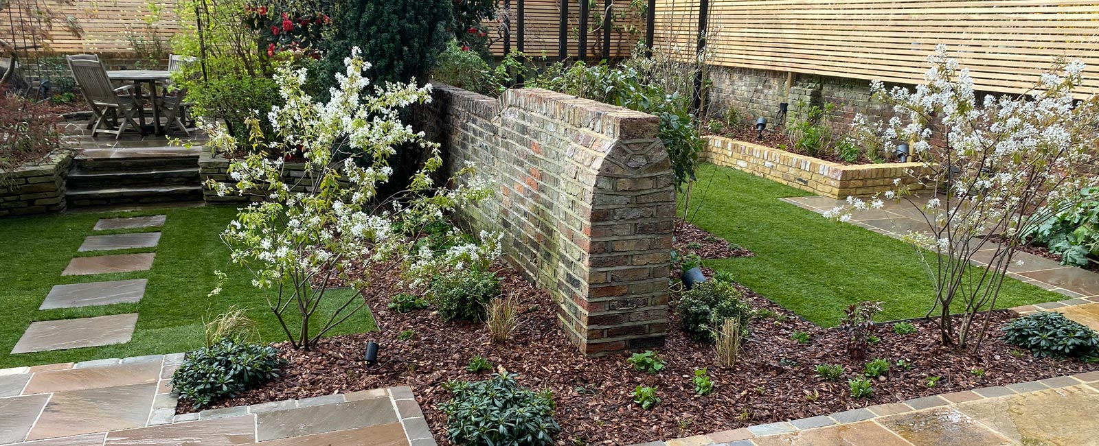 With low-maintenance shrubs, natural materials, sandstone paving, larch screening and complementary brickwork, two spaces work independently or as one larger area for relaxation and entertainment.