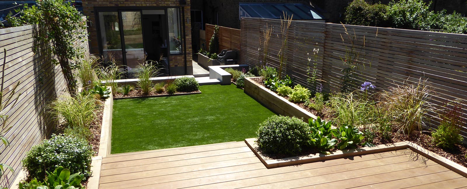 View of the garden looking across from the raised decking area, across the lawn, to the dining/seating area on the patio to the rear of the house.