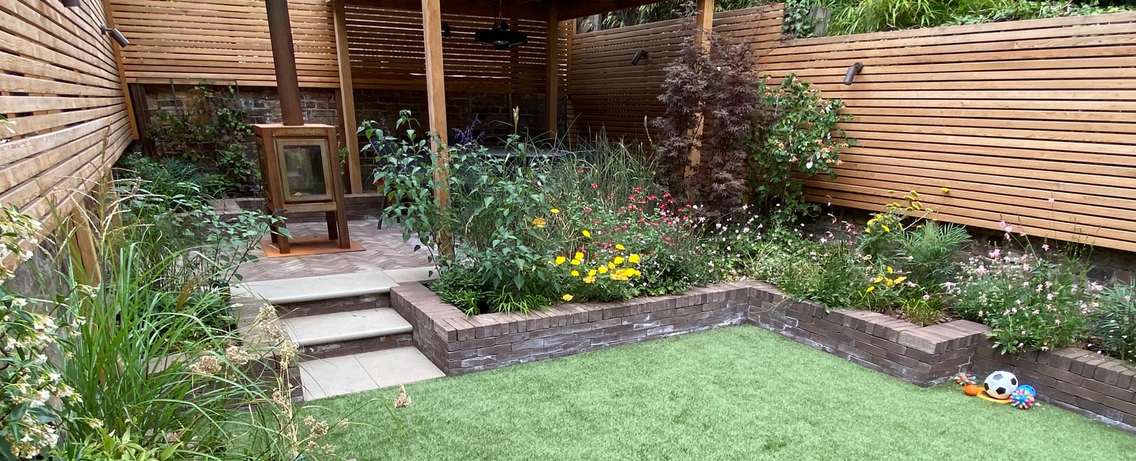 The bespoke cedar pergola is complemented by larch screening, sepia brick pavers and beds, grey sandstone steps, and planting of shrubs, grasses, climbers and perennials.