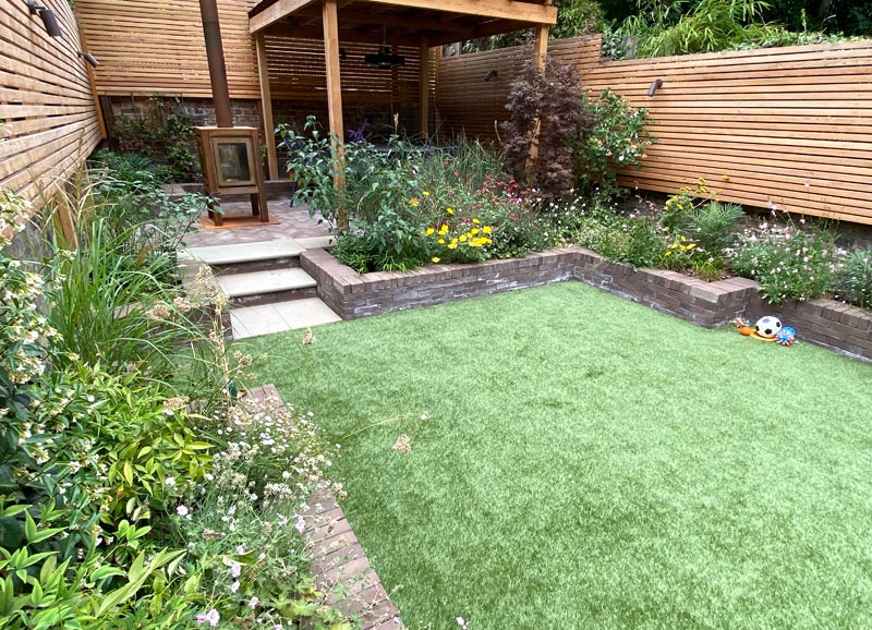 The bespoke cedar pergola is complemented by larch screening, sepia brick pavers and beds, grey sandstone steps, and planting of shrubs, grasses, climbers and perennials.