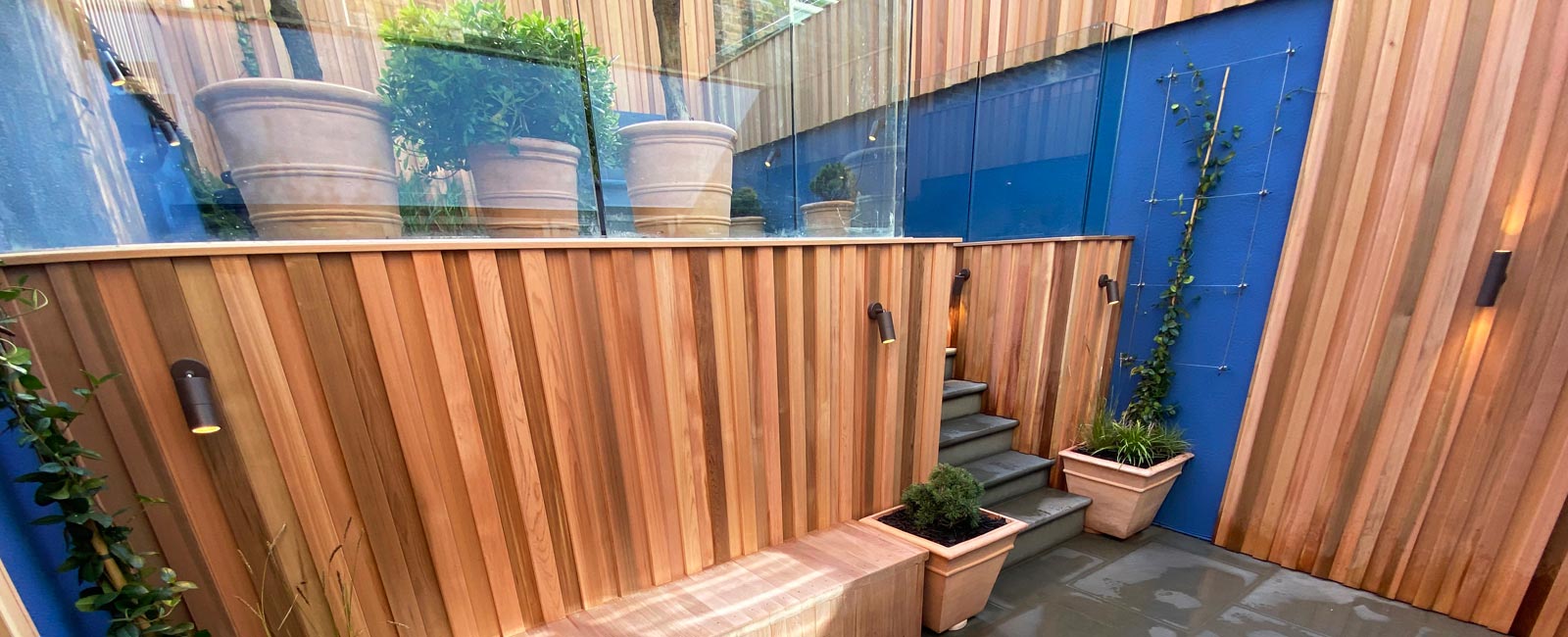 Features include cedar cladding, painted walls, grey sandstone steps, spotlights, a zinc water feature, large pots, space for seating and a fire pit, plus a variety of trees, shrubs, grasses, ferns and climbers.