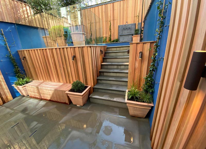 Features include cedar cladding, painted walls, grey sandstone steps, spotlights, a zinc water feature, large pots, space for seating and a fire pit, plus a variety of trees, shrubs, grasses, ferns and climbers.